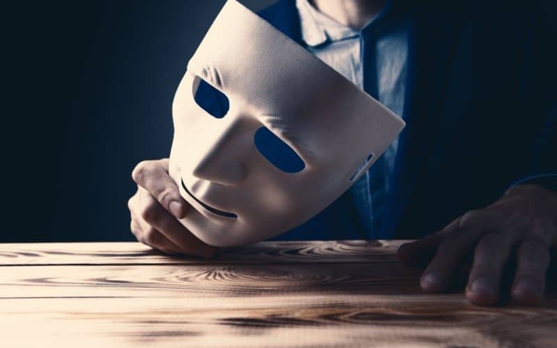 A man in a suit holds a mask above a wooden table, symbolising a training certification or role-playing scenario.